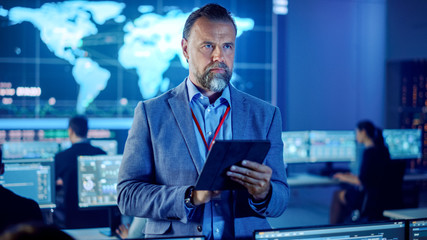 Fototapeta premium Male Project Leader is Standing with Tablet Computer and Making Mental Calculations. Data Science Engineers Work Around Him. Telecommunications Control Monitoring Room with a Global Map on Big Screen.
