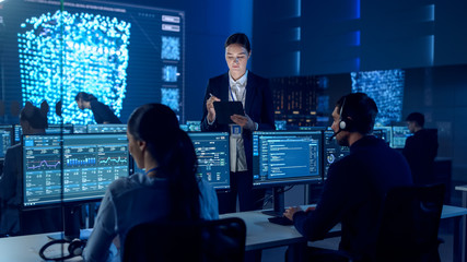 Female Project Leader is Standing with Tablet Computer and Checking Work Data. Science Engineers Work Around Her. Telecommunications Control Monitoring Room with Neural Network on Big Screen.