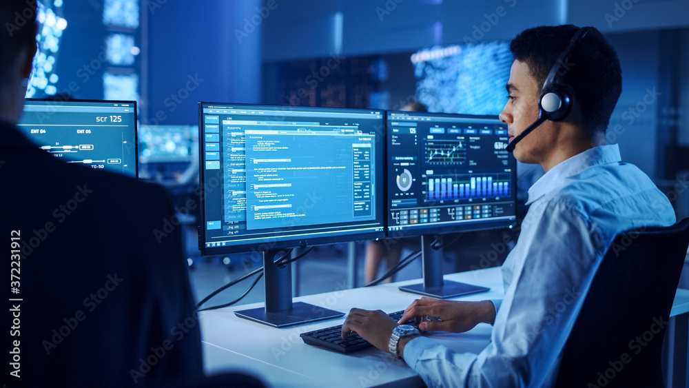 Poster Confident Male Data Scientist Works on Personal Computer Wearing a Headset in Big Infrastructure Control and Monitoring Room. Young Engineer in a Call Center Office Room with Colleagues.