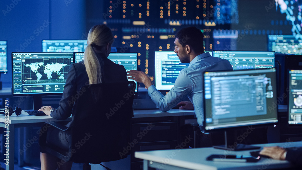 Aufkleber Confident Female Data Scientist Works Closely to her Young Colleague in Big Control and Monitoring Room with Neural Network. Young Specialists Talk and Discuss Work in a Room with Other Employees.