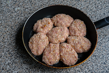 Cutlets, made from raw minced meat, lie tightly in a frying pan before frying, which sits on a grayish spotty countertop.