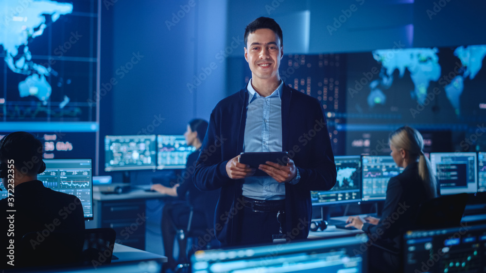 Poster Confident Successful Male Project Leader in a Computer Science Engineer Office Standing with Tablet Computer. He Looks at Camera and Smiles. Control Monitoring Room with a Global Map on Big Screen.