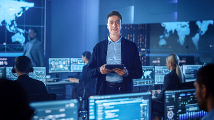 Confident Successful Male Project Leader in a Computer Science Engineer Office Standing with Tablet Computer. He Looks at Camera and Smiles. Control Monitoring Room with a Global Map on Big Screen.