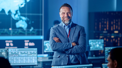 Fototapeta premium Confident Successful Senior Male Project Leader in a Computer Science Engineer Office Standing with Crossed Arms. He Looks at Camera and Smiles. Control Monitoring Room with a Global Map on Big Screen