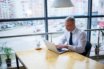 Male freelancer is working in a cafe on a new business project. Sits at a large window at the table. Looks at a laptop screen with a cup of coffee