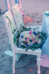 Elegant maritime wedding decoration on the beach, boho white chair with pastell green covers, beautiful big bride bouquet, pink, blue, yellow roses and greenery in sand background. Sea ceremony.