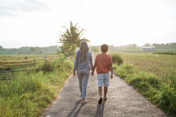 view from behind veiled mother and her son walk away from the road in the middle of rice field at...