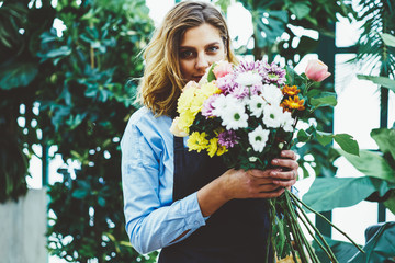 Half length portrait of positive female florist in apron looking at camera and holding bouquet with beautiful flowers, attractive caucasian saleswoman enjoying of time for hobby in green shop