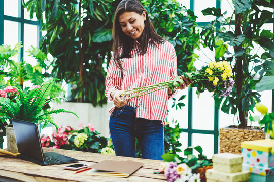 Cheerful asian woman designing floral composition while working in glasshouse near desk, successful female owner of flower shop making blooming bouquet with astera satisfied with job in orangery