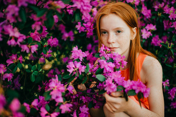 Expressive young girl with long red hair and pale skin sniffs flowers in the garden