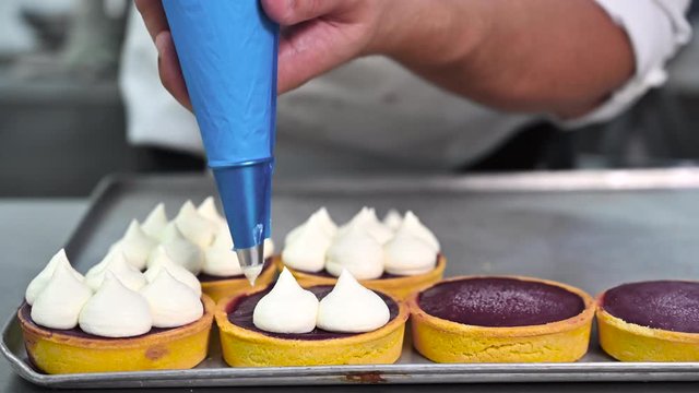 Pastry chef decorates biscuit tartlet with cream from pastry bag, close-up. Preparation of cake at commercial bakery with piping bag. High quality 4k footage.