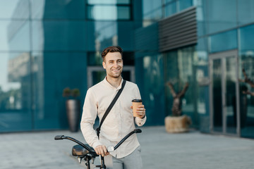 Coffee takeaway and eco transport. Smiling man holding glass and go with bicycle near office building