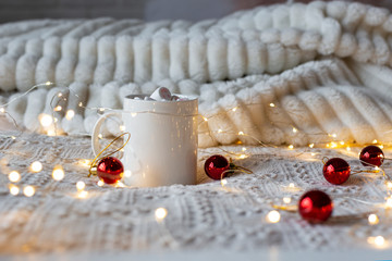 Christmas or New Year winter hot chocolate with marshmallows, festive light garland and red Christmas tree toys on a white bed, selective focus.