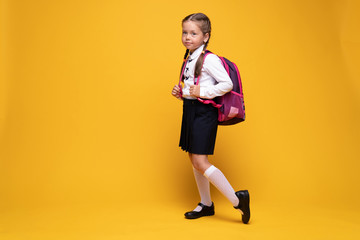 smiling little cute school girl in uniform with school bag, backpack, isolated on yellow background
