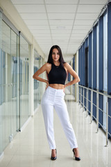 Fototapeta premium Young brunette woman, wearing white pants and black top, standing in light passageway with huge windows, posing with arms on her waist. Businesswoman on a break. Female portrait photography.
