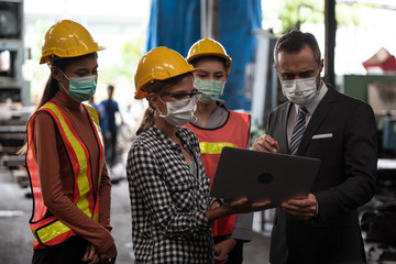 Industrial manager and women technician workers with Face mask,  hardhat. They walk together and using computer notebook for discussing manufacturing working plan in industrial Factory.