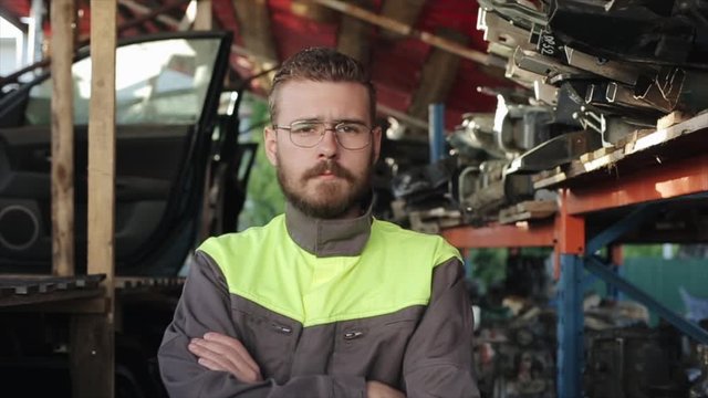 A serious young bearded auto mechanic in uniform stands with his arms crossed over his chest on the background of shelves with spare parts. Close-up