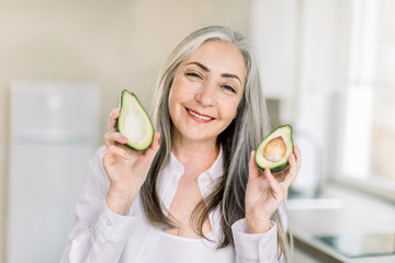 Health eating and lifestyle concept. Close up horizontal shot of a cheerful elderly woman with gray long hair, smiling to camera and holding avocado halves while standing in the kitchen