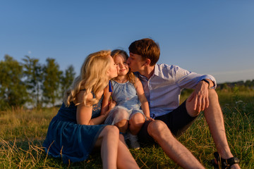 Dad mom and daughter are sitting together on the grass. Parents kiss their daughter on the cheeks.