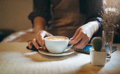 Close up of hand young asian  woman siting on chair and holding coffee cup to drink with small cactus in white pot and dry leaf in glass on table