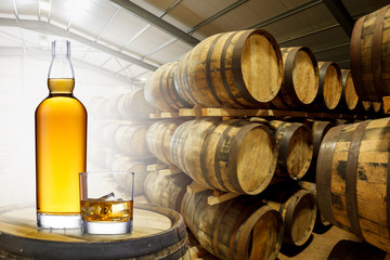 A bottle and glass of amber whisky set on top of an old barrel, in a barrel warehouse, back lit with light shinning through the open door.