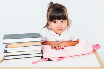 Brunette girl at school, sitting on a chair next to a table with books, leaning on her pink backpack on white background. School concept