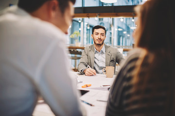 Portrait of young handsome bearded male manager of real estate agency sitting at table in front of colleagues during conference meeting making plans for trading strategy to increase incomes and sales