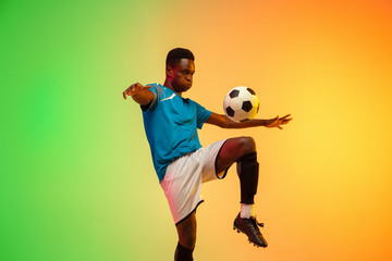 Unstoppable. African-american male soccer, football player training in action isolated on gradient studio background in neon light. Concept of motion, action, ahievements, healthy lifestyle. Youth