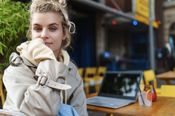 Close-up rear view of beatiful blond girl turn at camera while sitting coffee table with laptop computer and cocktail. Student studying outdoors. Female freelancer having break in cafe