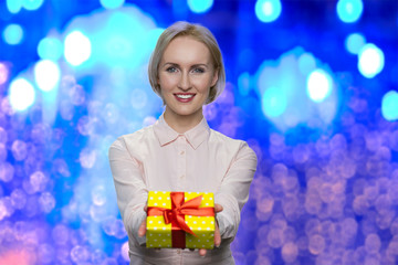 Pretty caucasian woman giving Christmas gift box. Young beautiful business woman handing present box on blue bokeh lights background. Special bonus offer.