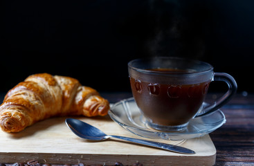 
Hot coffee in a cup placed on a wooden floor with a croissant placed behind it.