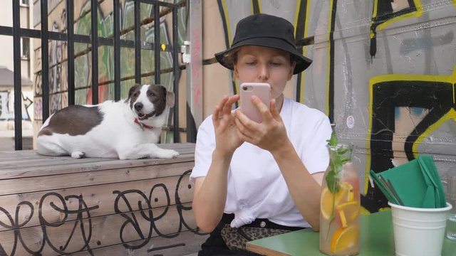 Girl in a bucket hat using her smartphone while waiting in a cafe with pretty dog chilling on the background. Graffiti walls. City life concept. 