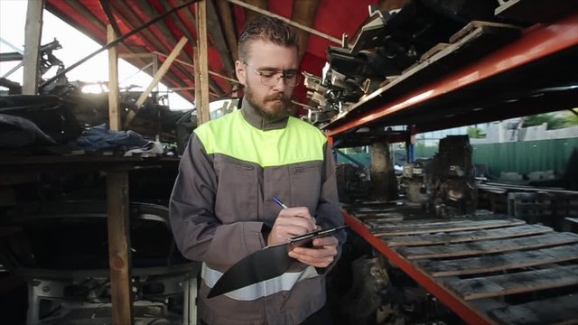 Young auto mechanic with a beard and glasses stands in a warehouse and takes notes in a notebook. The camera zooms in on a close-up. Slow motion