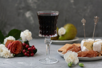 Crystal glass with red wine in the middle of the frame, next to a plate with ham and Camembert cheese and flowers. Composition on a gray concrete background.