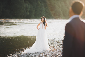 Fototapeta na wymiar Elegant stylish happy blonde bride and gorgeous groom on the background of a beautiful river in the mountains