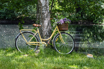 The old vintage yellow bicycle with lilac flowers in the basket stands by a pond in the parc in summer