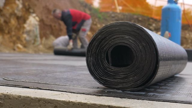SLOW MOTION, MACRO, DOF: Thick roll of tar lies on the construction site ground as unrecognizable contractors work in the blurry background. A crew of builders are waterproofing the house foundation.