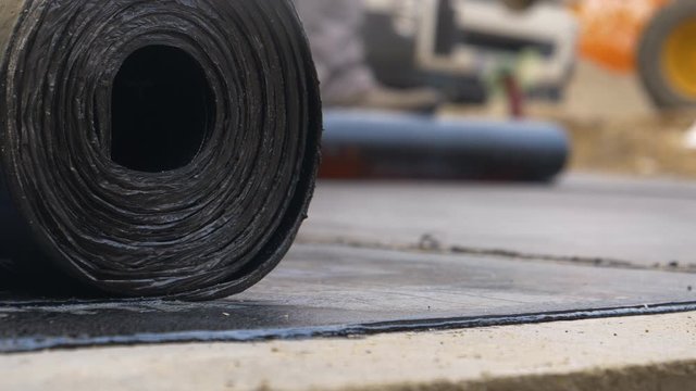 SLOW MOTION, MACRO, DOF: Detailed shot of bitumen roll as unrecognizable worker in the background burns another layer of hydroinsulation on the foundation of a house under construction. Waterproofing