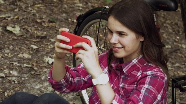 Young beautiful caucasian woman sits resting near the orange city rental bike and makes a photo on a smart phone. Smiling girl woman with mobile phone in hands sits on grass next to her bicycle