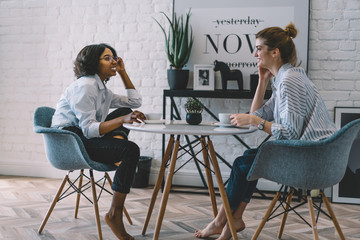 Fototapeta premium Happy two hipster girls communicating with each other drinking tasty coffee in stylish apartment.Positive african american female enjoying funny conversation with best friend in cozy room