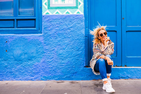 Chererful And Relaxed Beautiful Woman Sit Down Outside A Colorful Blue House In The Street - Home Properties Owner People Concept - Travel Lifestyle And Scenic Places To Discover