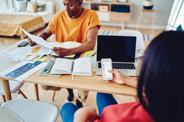 Dark skinned colleagues working together at office using modern technologies, cropped image of african american woman holding smartphone for networking and chatting sitting at desktop with laptop.