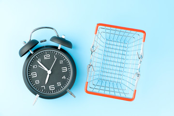 Black vintage analog alarm clock and small shopping basket on blue background.