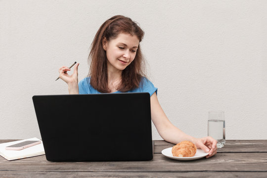 Young woman has distance work, online meeting, video call, zoom conference on a laptop sitting at a wooden table on a light background and thinking about something looking at a plate with a croissant