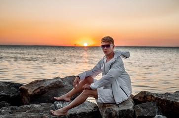 a guy in white clothes and sunglasses sitting on the stones near the sea at sunset