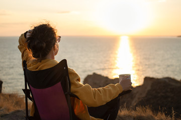 Fototapeta premium Back view of young woman traveler holding iron mug cup with tea or coffee,enjoying sunset scenery view of the sea landscape, sitting in touristic chair. Travel camping and adventure lifestyle concept