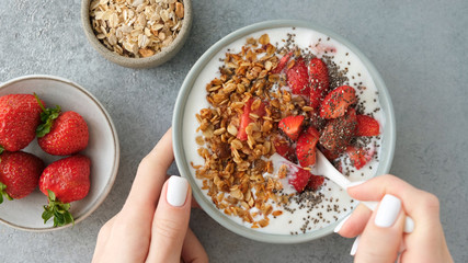 Eating granola with strawberries and yogurt. Female hands holding bowl of homemade honey oat granola with strawberries and chia seeds. Top view