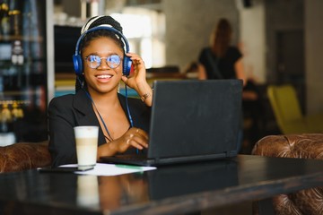 Happy black girl in wireless headphones studying online, using laptop and taking notes, cafe interior, copy space