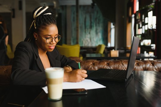 Student girl browsing Internet, using free wi-fi at cafe. African freelancer thinking on ideas for her blog, using laptop at co-working space, resting hand on wooden table, looking with inspired smile
