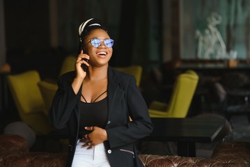 Smiling dark skinned woman in trendy wear laving cafe talking on mobile phone via roaming tariffs,african american millennial female walking and making smartphone call satisfied with connection
